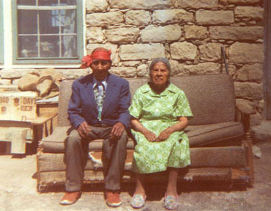 Adopted Hopi Grandparents sitting side by side on couch outside their Adobe dwelling.